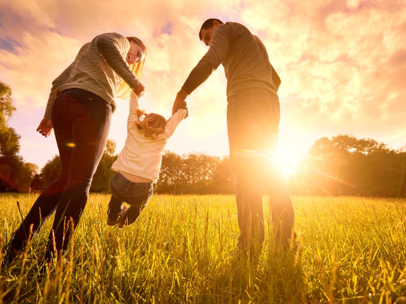 Parents holding a baby's hands while walking in a field at evening