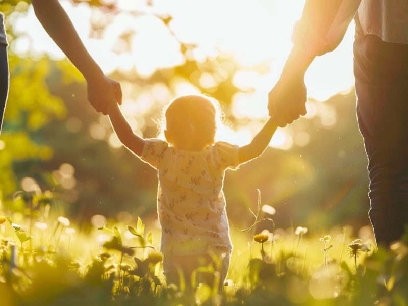 Two parents holding a baby's hands while walking in a field